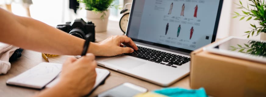Zakupy online, osoba przegląda ubrania na laptopie, notatnik i aparat na stole.