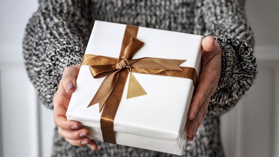 Biały prezent z brązową wstążką i złotą ozdobą, trzymany w dłoniach.