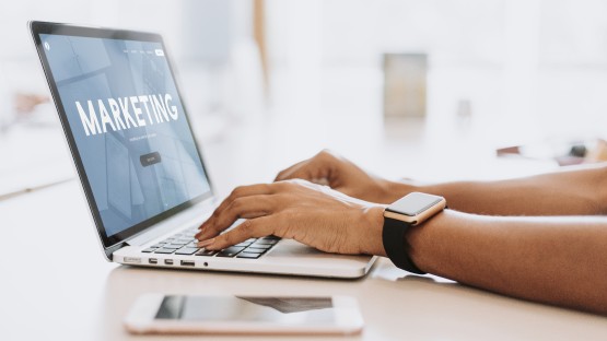 Osoba korzystająca z laptopa z wyświetlonym napisem "MARKETING", obok leży smartfon.